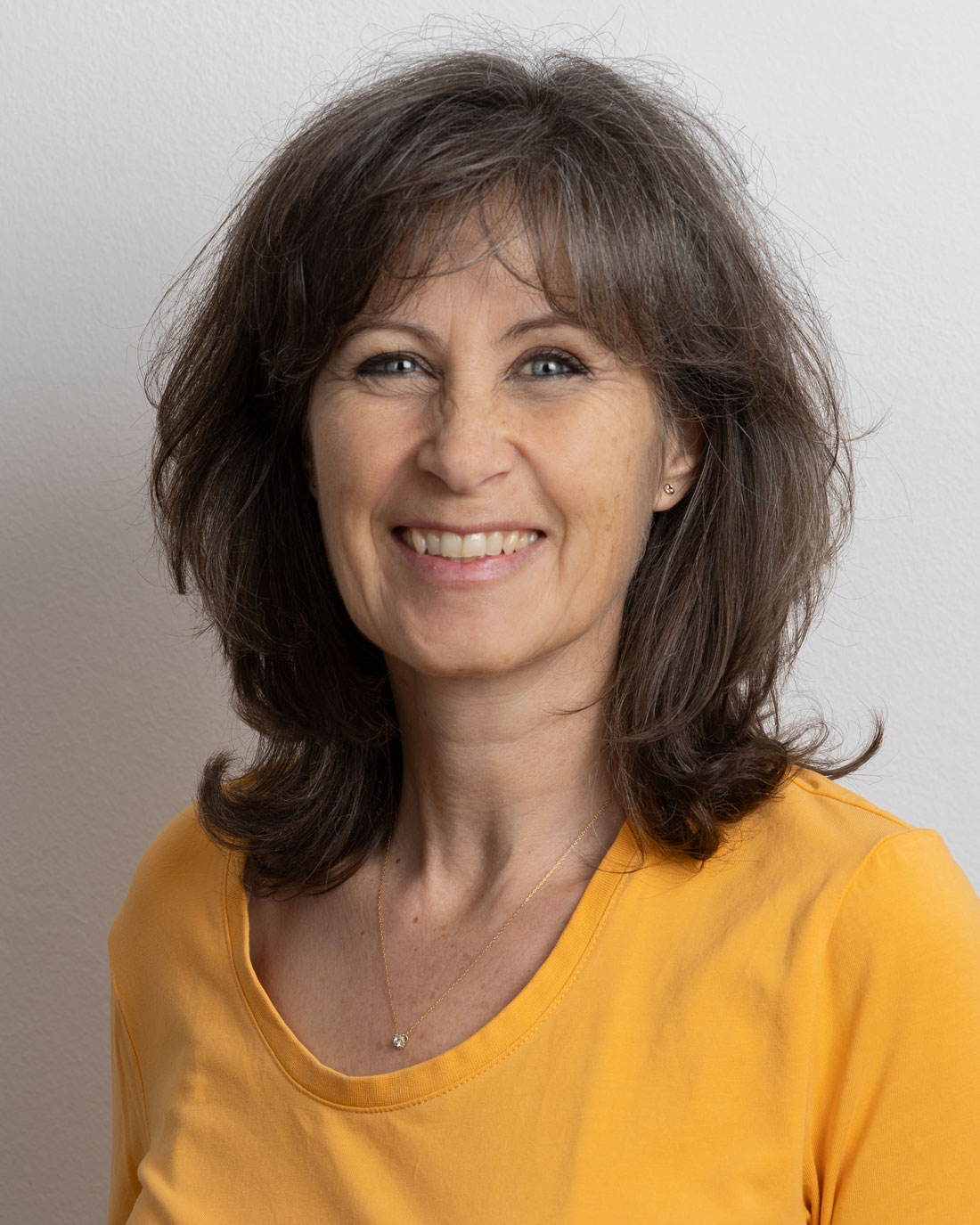 Ein Nahaufnahme-Porträt von Andrea Engel, die herzlich lächelt, ein gelbes Shirt und eine zarte Halskette trägt. Ihr Haar ist mittellang, braun und wellig, und sie hat einen freundlichen und zugänglichen Ausdruck.